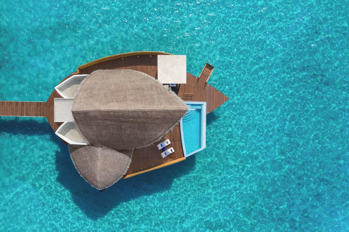 Un bungalow sur pilotis avec un grand rocher servant d’élément central, une eau d’un bleu cristallin et une terrasse en bois.