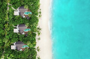 Vista aérea de um resort  à beira-mar com várias bangalôs sobre a água, uma praia arenosa e águas azul-turquesa.