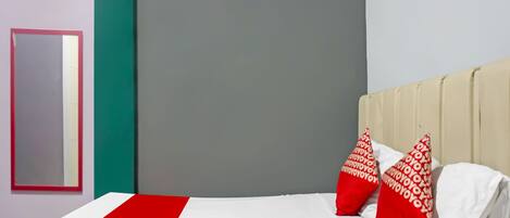A hotel room with a bed, red and white pillows, a red and white striped area rug, and a grey wall with a mirror.
