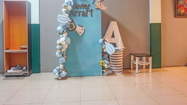 A room with a blue door decorated with balloons and a name sign. There is a bookshelf with books and a pillow on a shelf. A striped trash can and a small table with a chair are also visible. - Hotel O Puspita Guesthouse (Bekasi)
