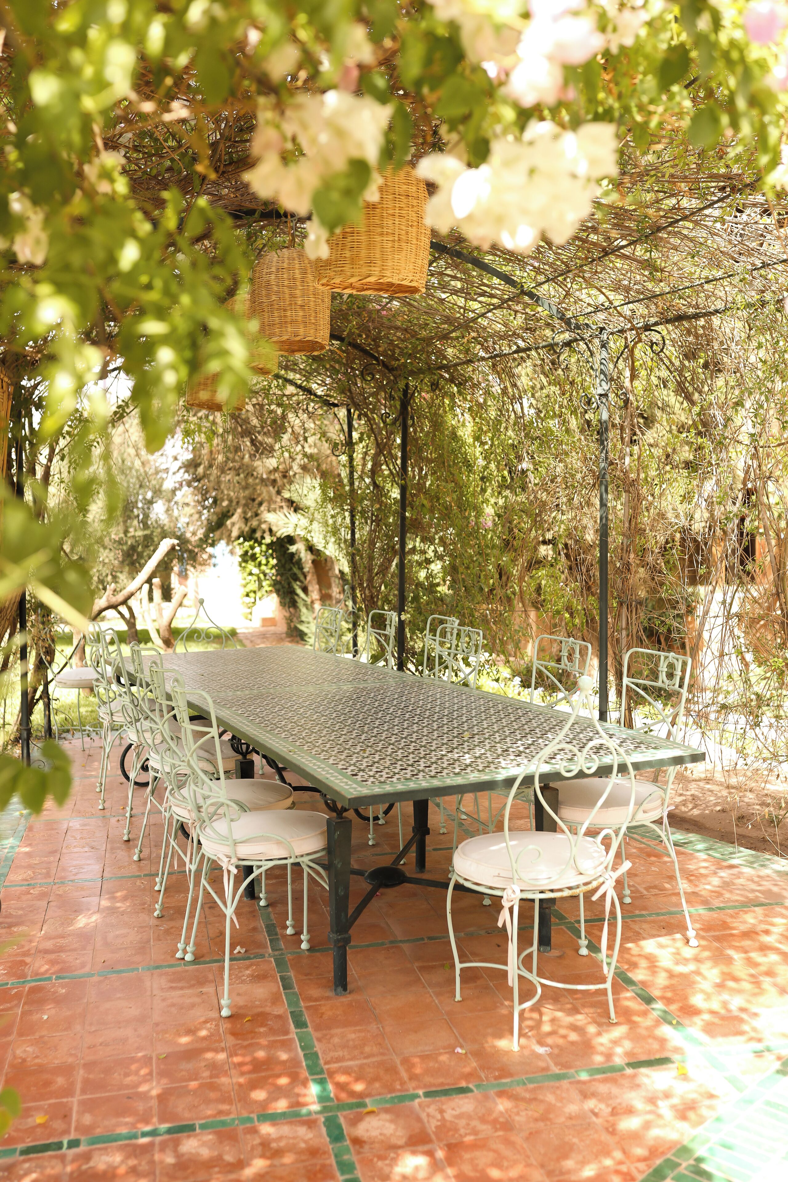 Un espacio de comedor al aire libre con una mesa y sillas de metal, rodeado de follaje y una pérgola.