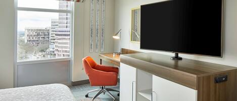 A hotel room with a bed, a desk with a computer, a TV, and a window with a city view.