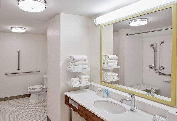 Un baño moderno con lavamanos, espejo y retrete blancos. Toallas blancas apiladas en un estante, y una barra de apoyo junto al inodoro.