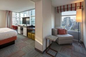 A hotel room with a bed, a desk with a computer, a chair, a sofa, and a large window offering a city view.