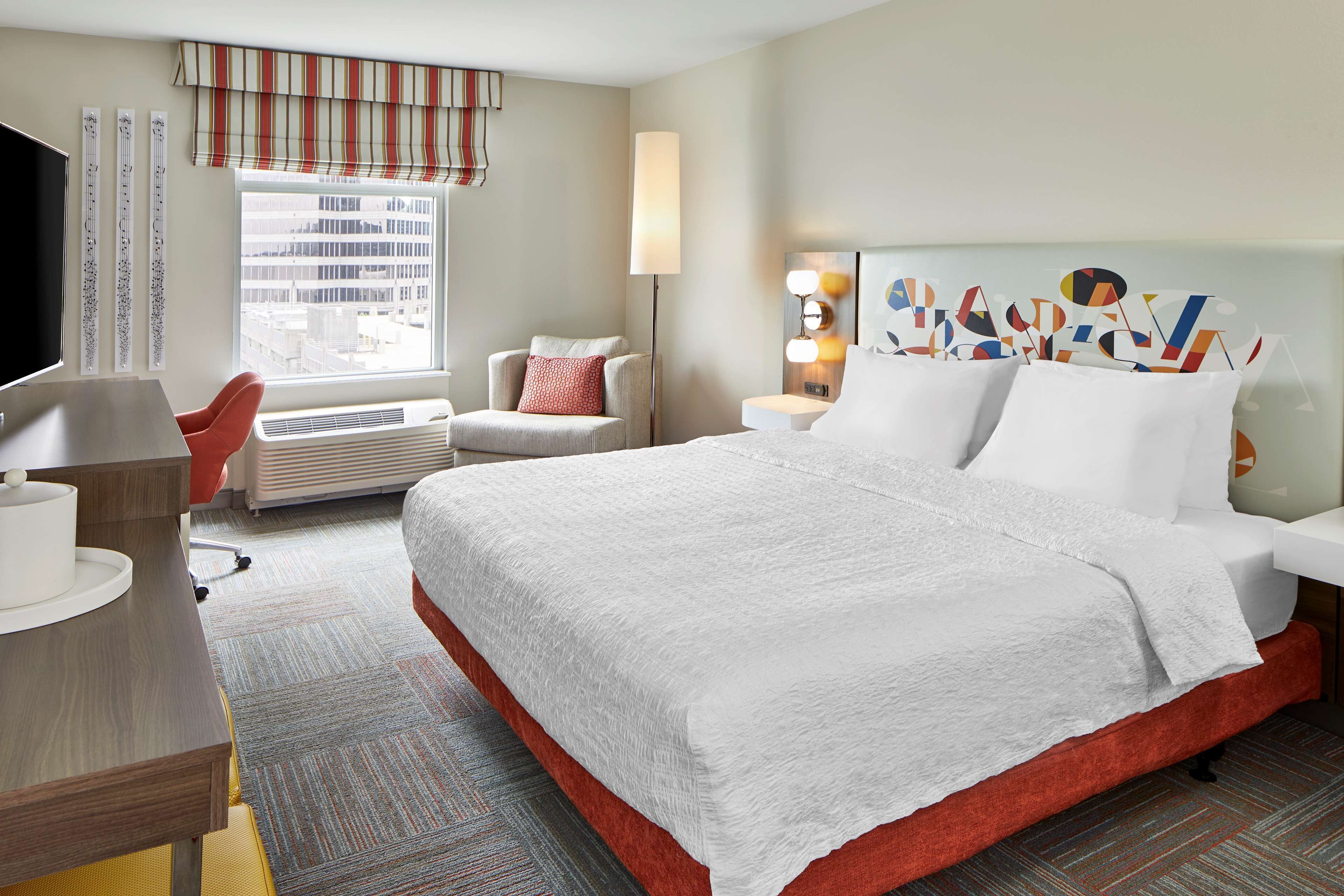 Habitación de hotel con una cama grande, un escritorio con silla, un televisor, una ventana con vistas a edificios y una pared decorada con una obra de arte colorida.