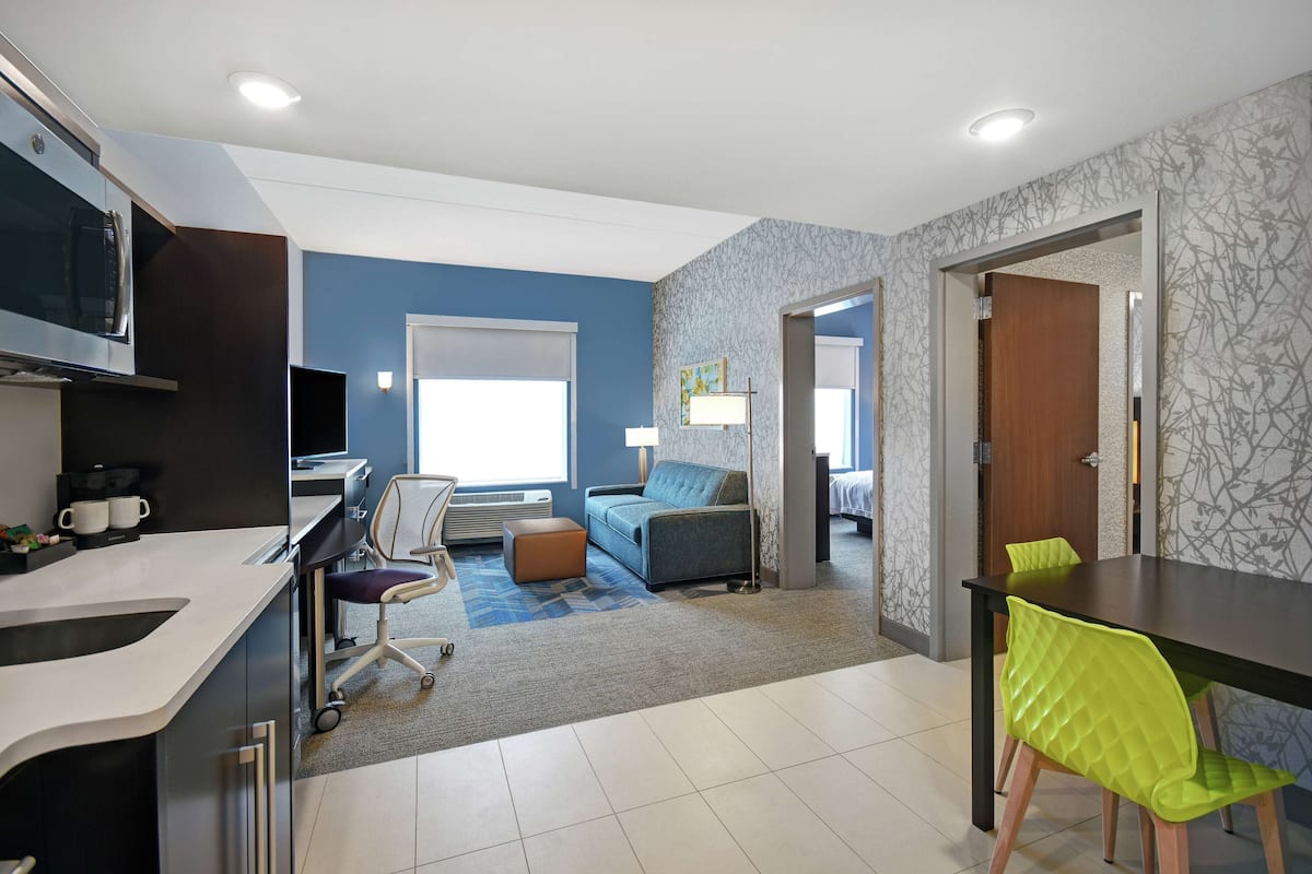 Ein modernes Hotelzimmer mit Küchenzeile, einem Esstisch, einer Couch und einem durch eine offene Tür einsehbaren separaten Schlafzimmer.