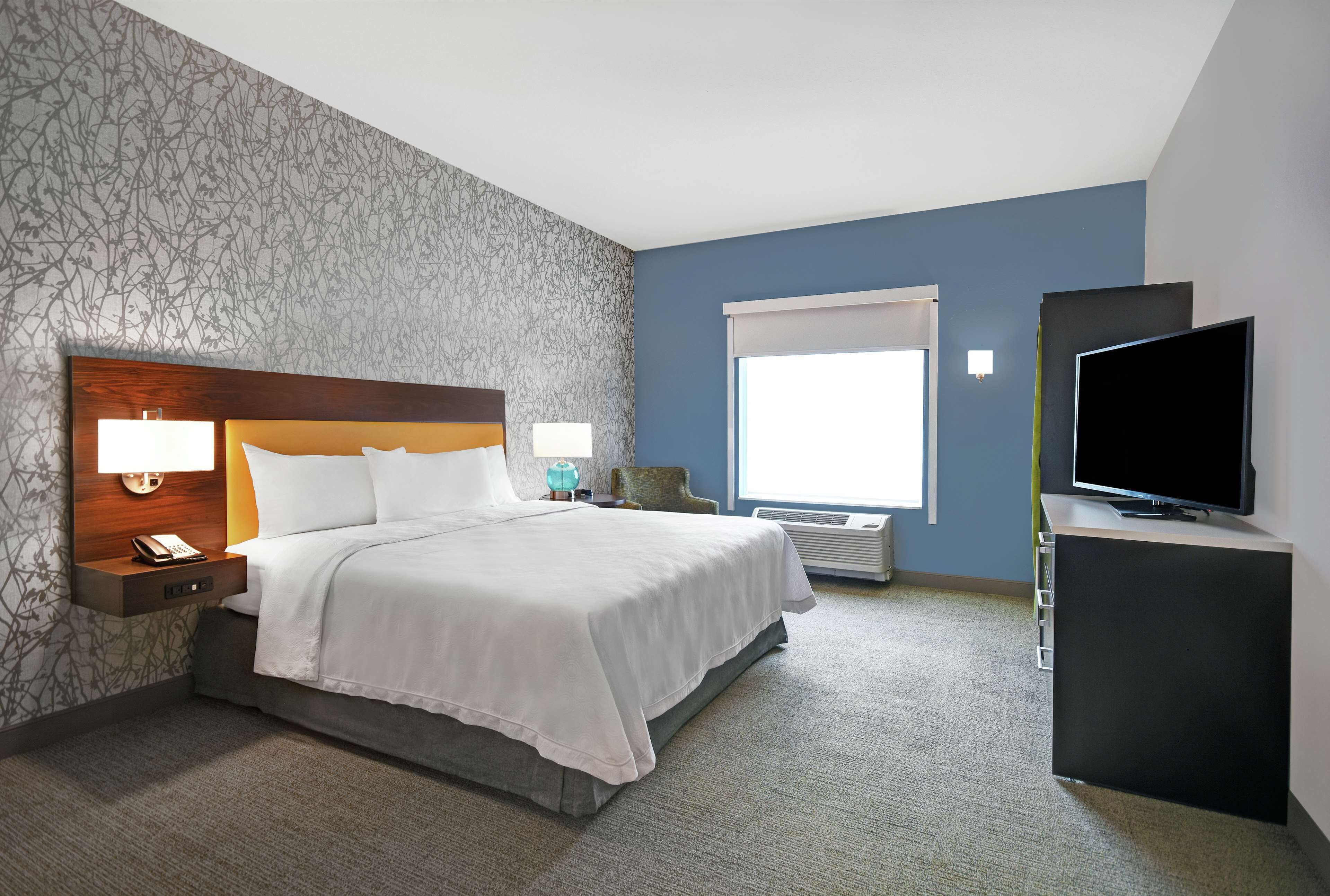A hotel room with a large bed, a flat-screen TV, a nightstand with a lamp, and a window with blinds.
