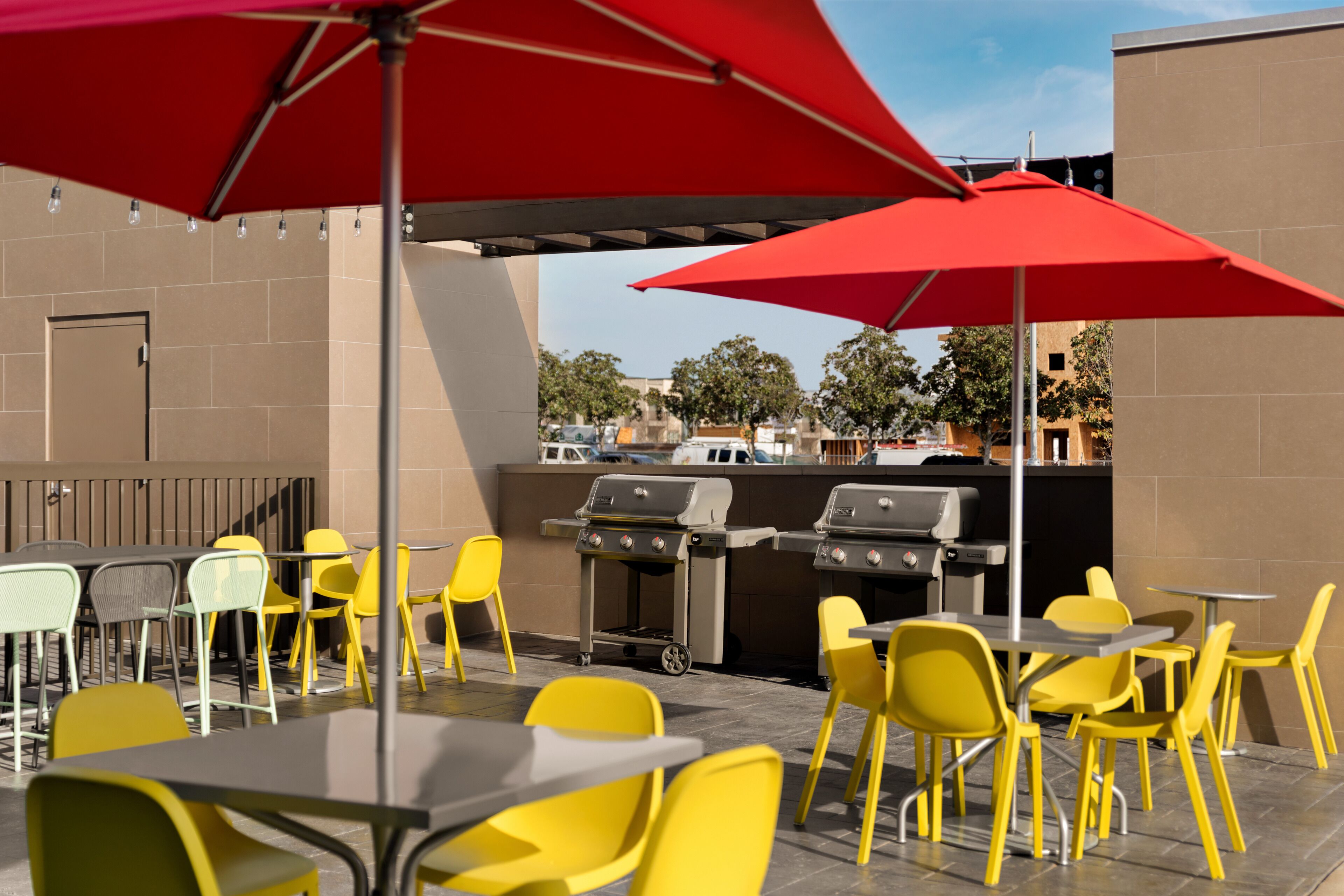 An outdoor patio with yellow chairs and tables, red umbrellas, and a barbecue grill.