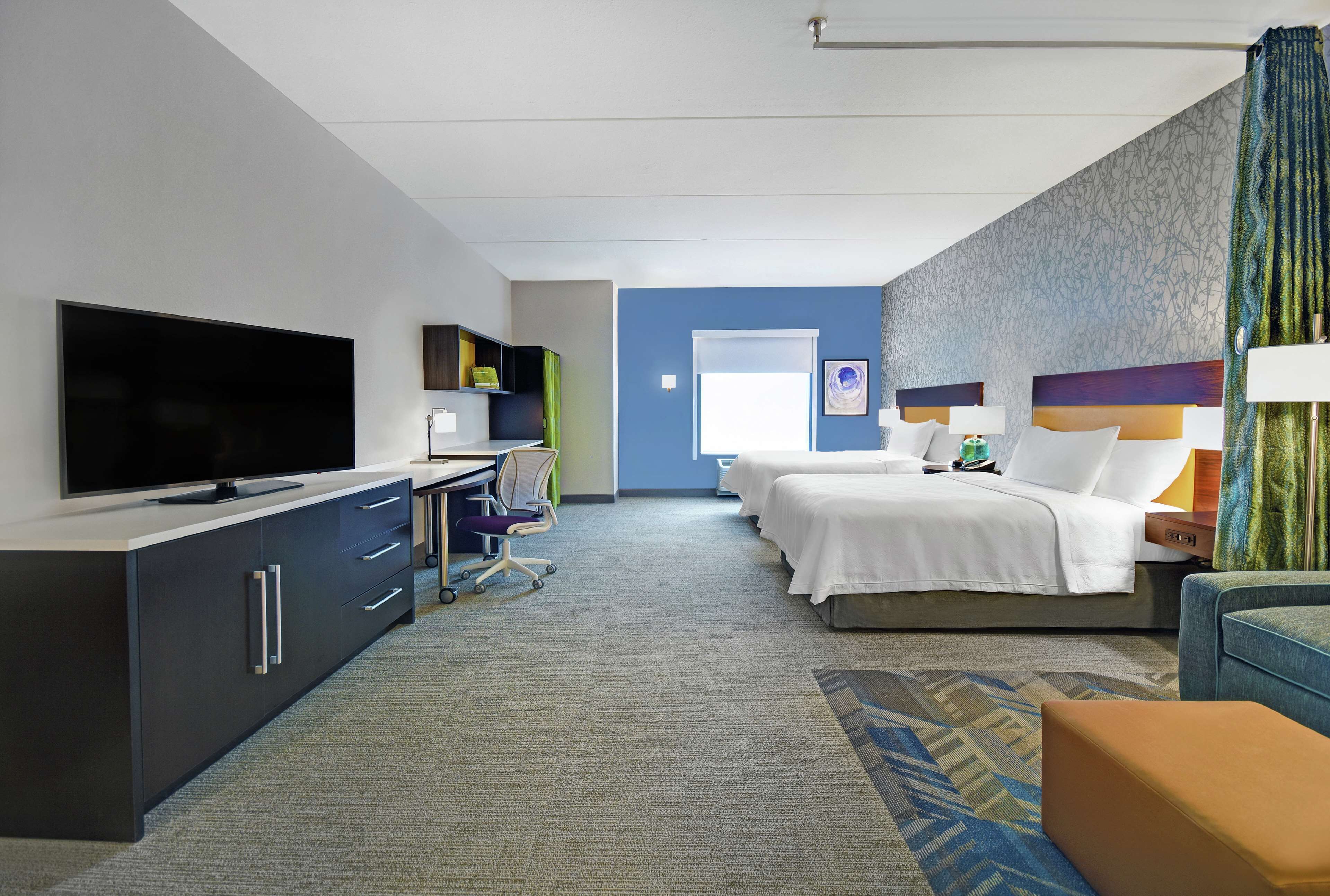 A hotel room with a flat-screen TV, a desk with a chair, two beds, a sofa, and a patterned carpet.