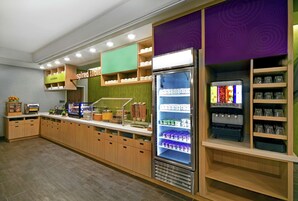 Zona de desayuno autoservicio con mostrador refrigerado, estantes con tazas y recipientes, y máquina de café.