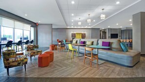 A spacious lounge area with a sofa, chairs, and tables, featuring a large window and a modern ceiling design. - Home2 Suites by Hilton Lawrenceville Atlanta Sugarloaf, GA (Lawrenceville)