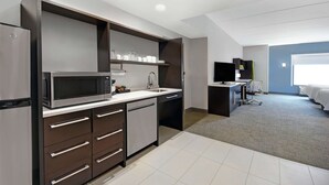A modern kitchen with a microwave, refrigerator, and built-in oven. There is a sink and a countertop with a chair. The room has a gray carpet and white walls. - Home2 Suites by Hilton Lawrenceville Atlanta Sugarloaf, GA (Lawrenceville)