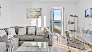 A modern living room with a grey sofa, a glass coffee table, and a view of the sea through an open door.