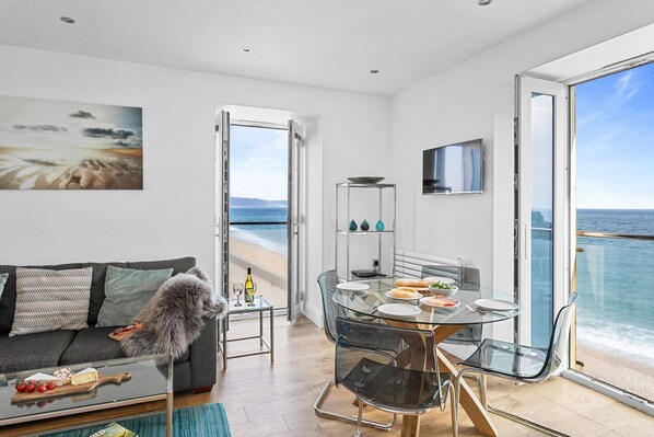 A modern living room with a glass dining table, a sofa, and a view of the sea. - 14 At The Beach, Torcross -  an apartment that sleeps 4 guests  in 2 bedrooms (Torcross)
