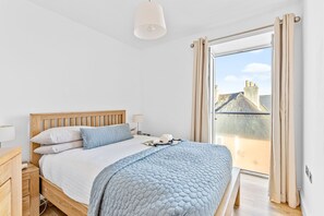 A bedroom with a wooden bed, a white ceiling lamp, and a view of a house through a large window. - 14 At The Beach, Torcross -  an apartment that sleeps 4 guests  in 2 bedrooms (Torcross)