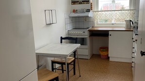 A compact kitchen with a refrigerator, stove, and a small dining table with two chairs. There is a window with a lace curtain and a view of buildings outside. - Four Bedroom Guesthouse in Fjerdingen, Harran (Fjerdingen)
