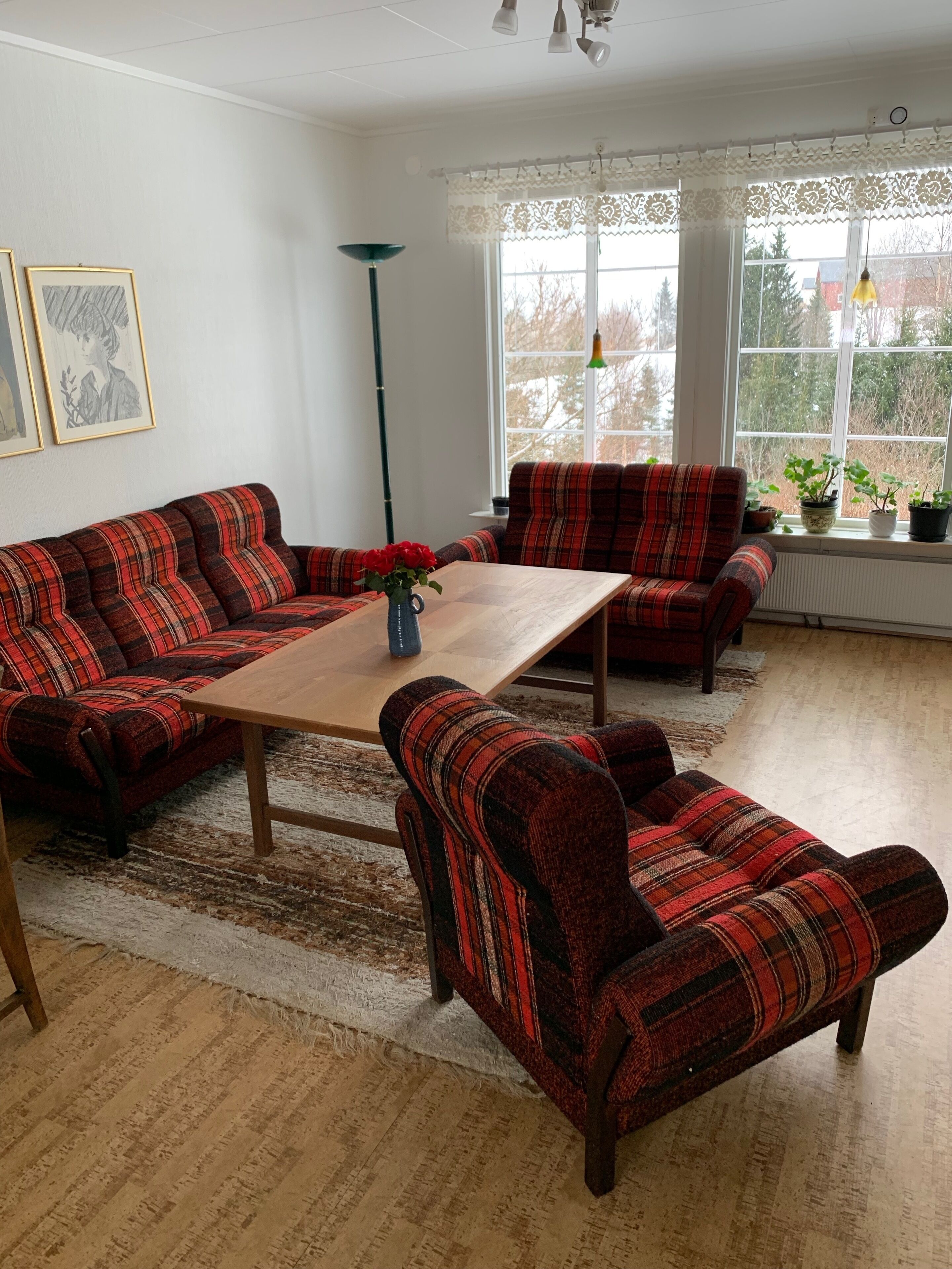 A living room with a red and black striped sofa, a wooden coffee table, and a matching armchair. There are two windows with lace curtains, a floor lamp, and a vase with red flowers on the coffee table.