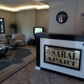 Uma área de recepção com uma placa que diz "SARAL APART" e uma televisão ao fundo.