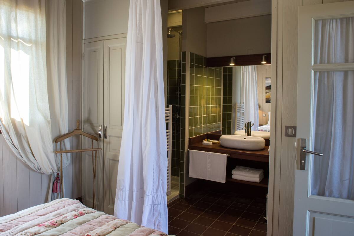 A bedroom with a bed, a hanging shower curtain, and a bathroom with a sink and mirror.