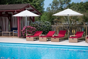 Un espace aménagé au bord de la piscine, avec des chaises longues et des parasols, une structure en bois et un arbuste aux fleurs roses.
