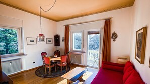 A cozy room with a dining table, chairs, a red sofa, and a view of trees through the windows. - Central, spacious and comfortable, easy to reach by car or train. (Merano)