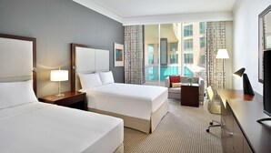 A hotel room with two beds, a desk, a chair, a TV, and a view of a swimming pool. - DoubleTree by Hilton Doha - Al Sadd (Doha)