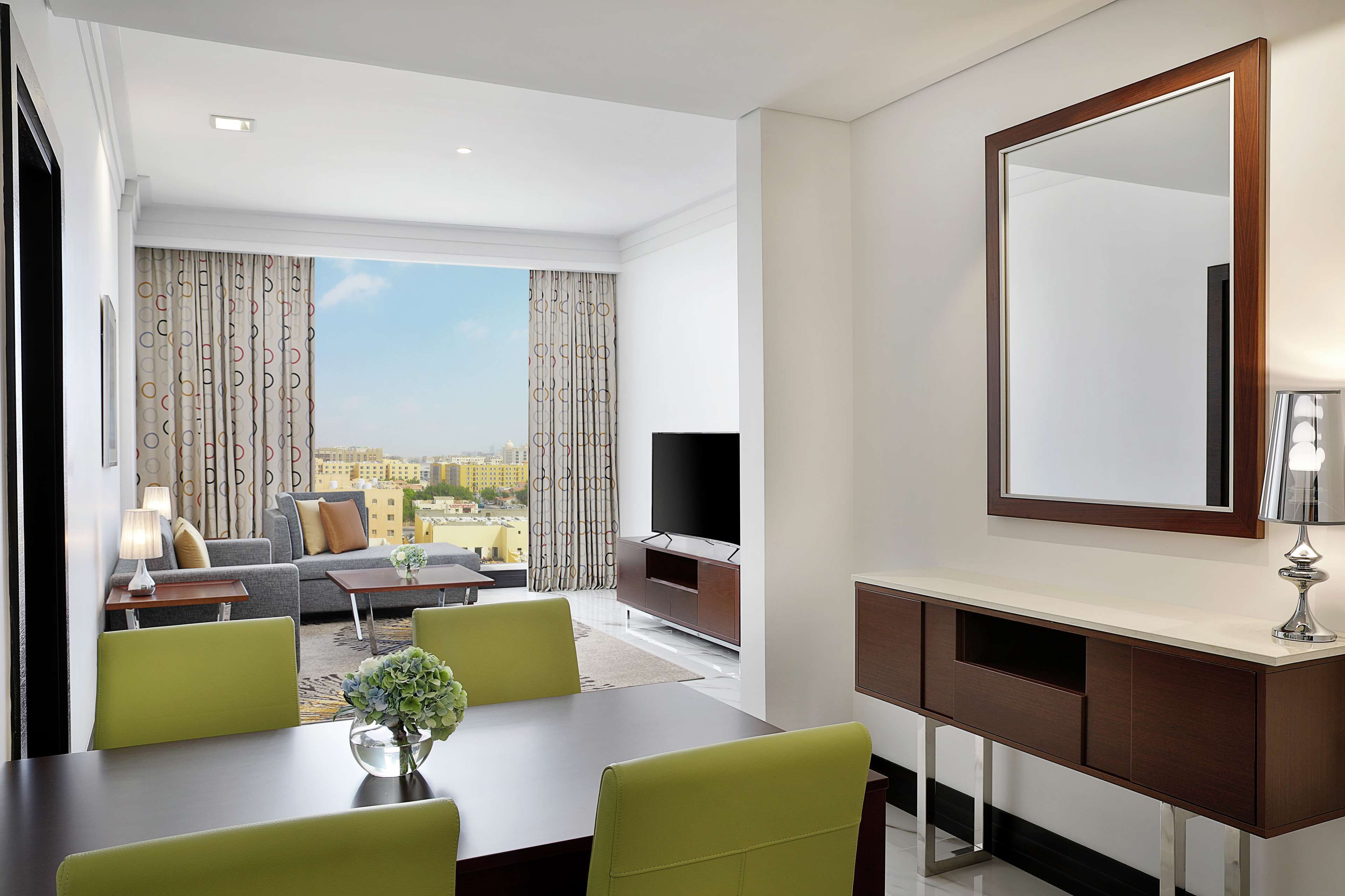 Una habitación de hotel moderna con comedor, sala de estar con sofá y televisor, y un amplio ventanal con vistas a la ciudad.