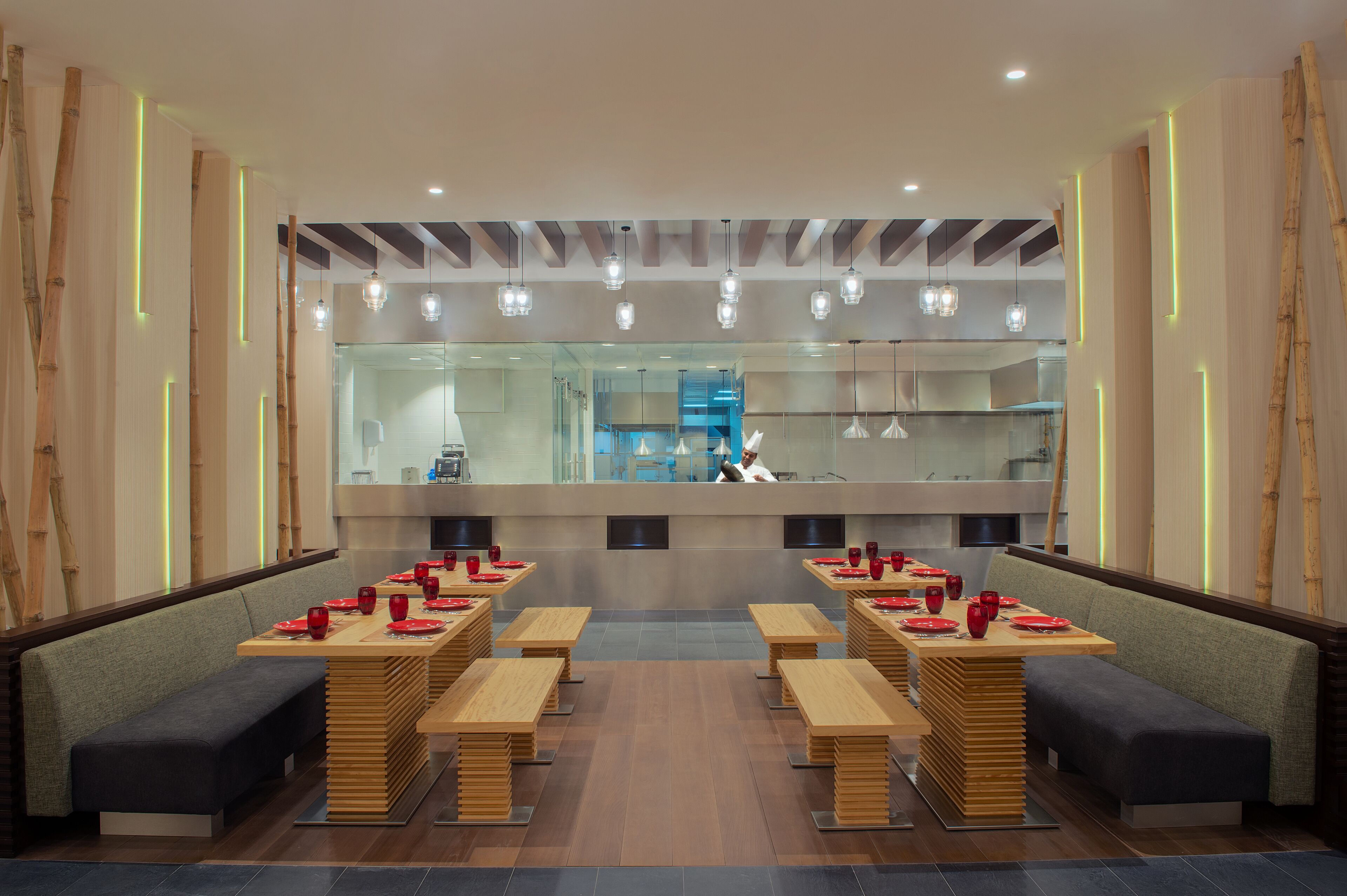 Un restaurante moderno con varias mesas y bancos de madera, un panel de vidrio que separa la zona de comedor de la cocina, y un diseño limpio y contemporáneo.
