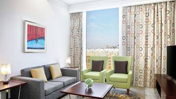 A modern hotel room with a grey sofa, two armchairs, a small table with a vase, a TV, and a painting on the wall.