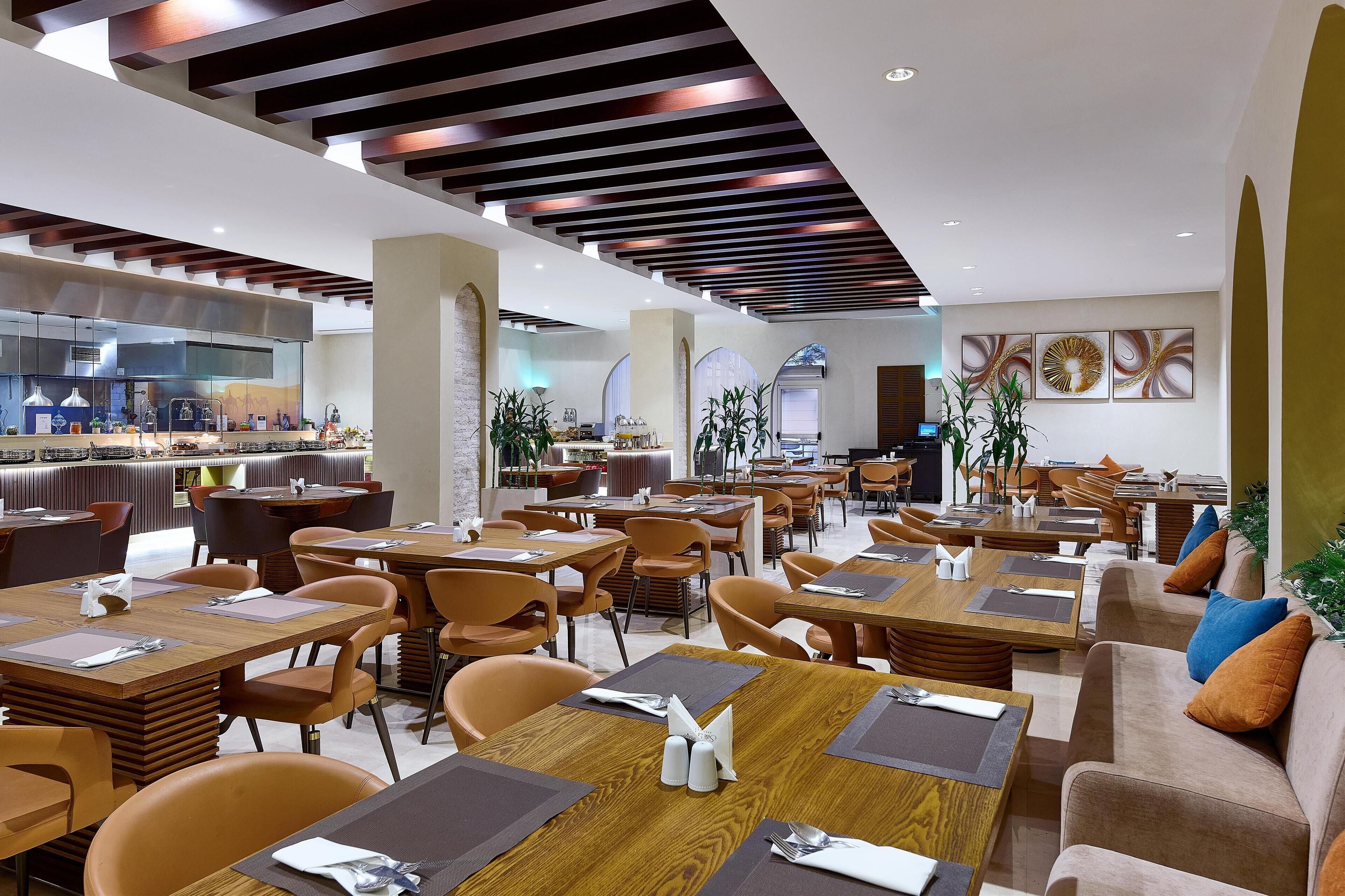 A modern restaurant interior with wooden tables and orange chairs, a kitchen area in the background, and decorative wall art.
