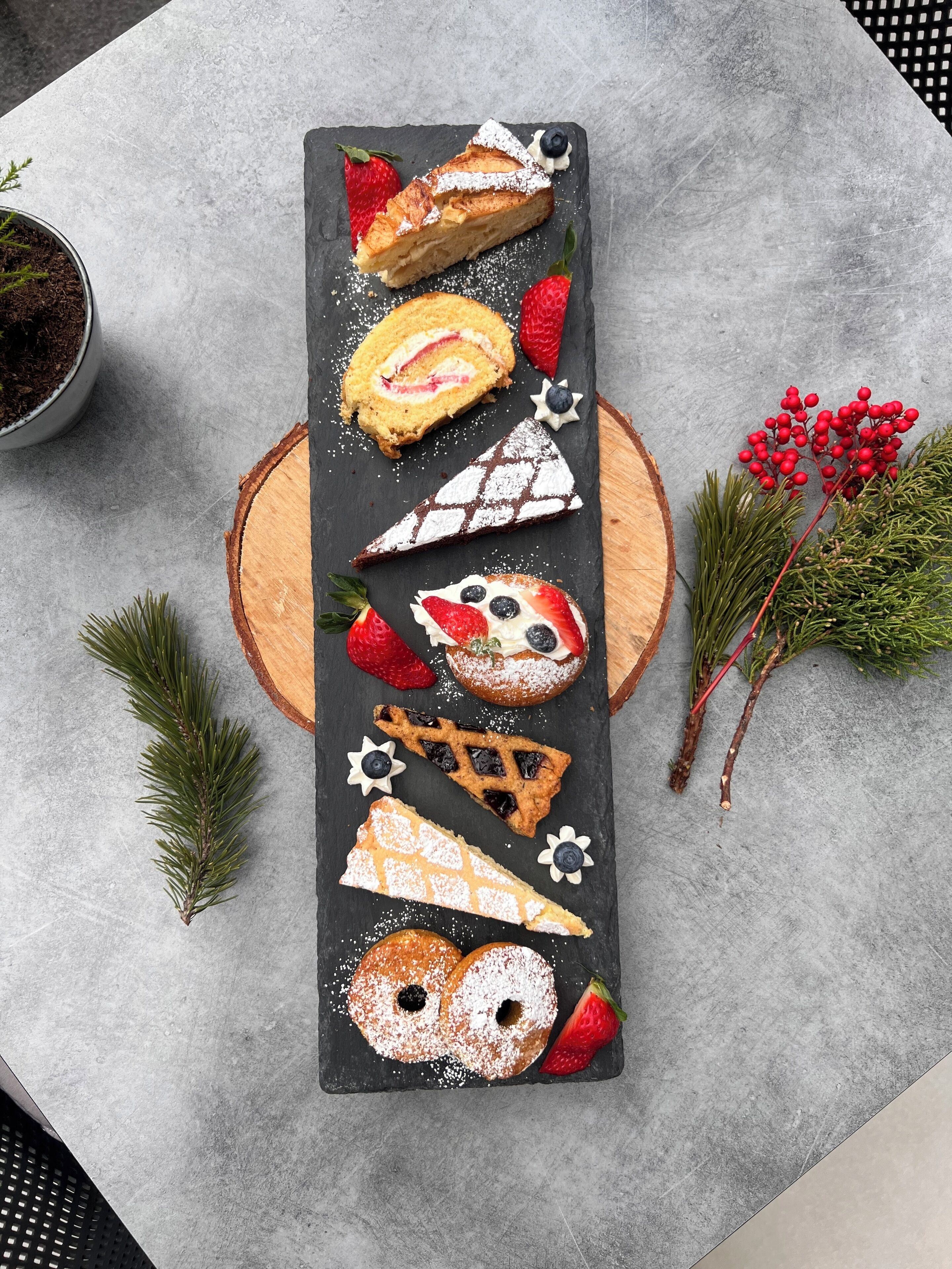 Une variété de pâtisseries et de desserts disposés sur une ardoise, accompagnés de fraises et de myrtilles fraîches, ainsi que de quelques éléments de décoration végétale.