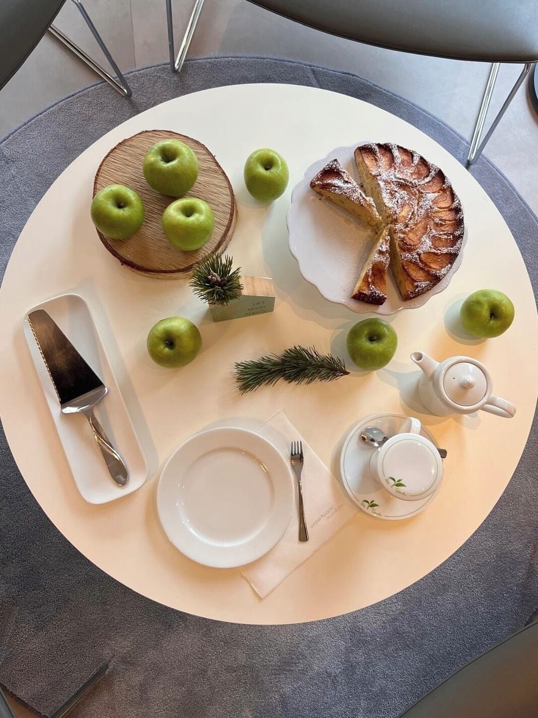 Una mesa redonda con un plato blanco, un cuchillo, un tenedor, una tetera y una taza. También hay varias manzanas verdes y una pequeña rama de pino sobre la mesa.