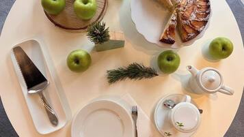 Una mesa redonda con un plato blanco, un cuchillo, un tenedor, una tetera y una taza. También hay varias manzanas verdes y una pequeña rama de pino sobre la mesa.