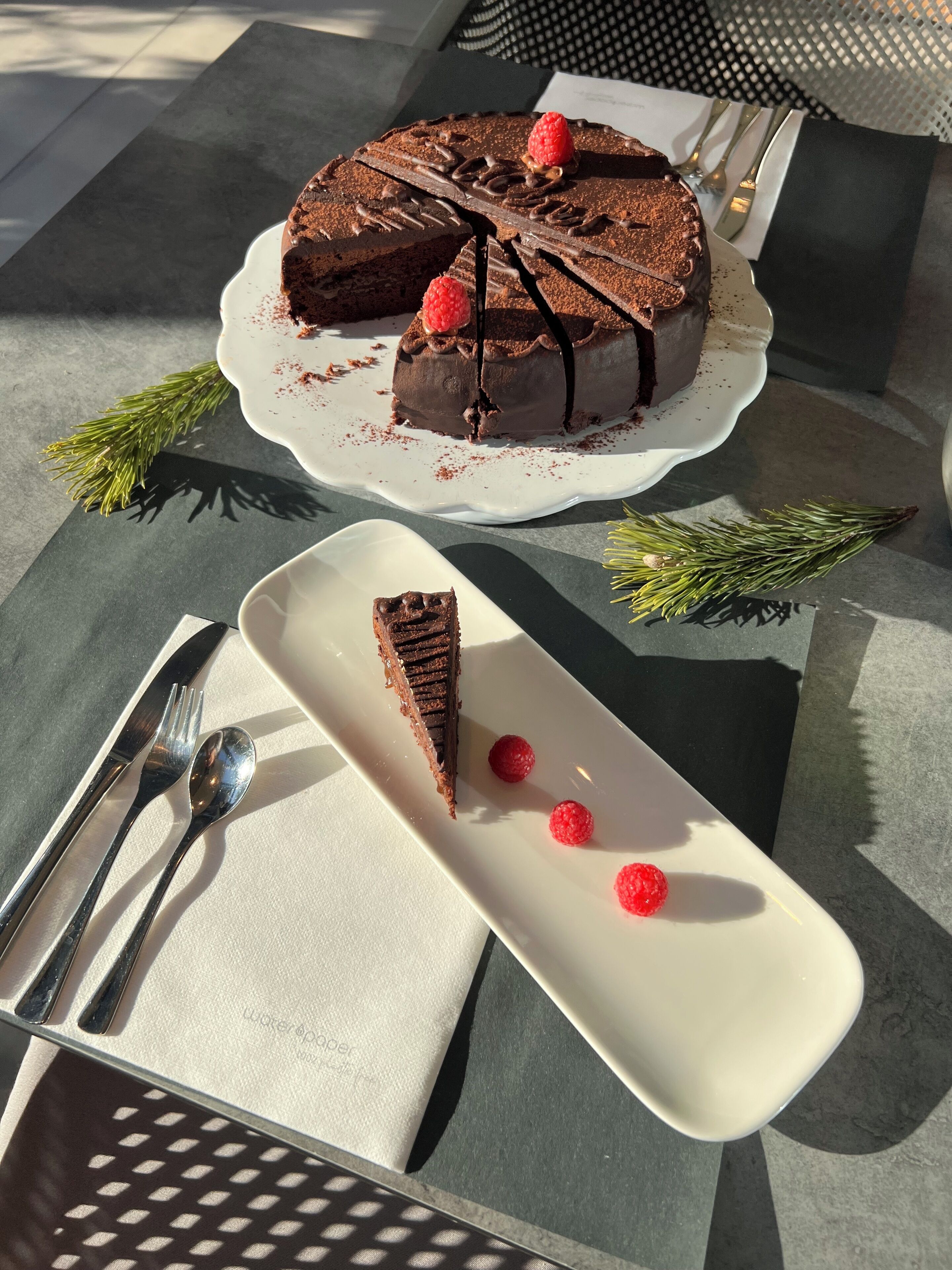 A chocolate cake with a slice cut out, placed on a white plate with a fork and spoon beside it.