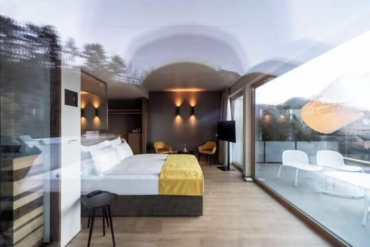 Une chambre moderne avec un grand lit, deux fauteuils, un téléviseur et un balcon agrémenté de chaises blanches.