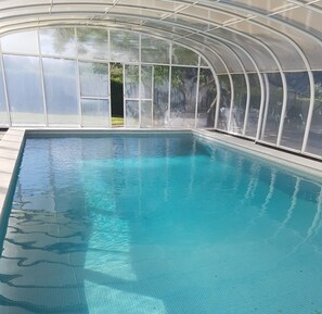 Une grande piscine intérieure à l’eau d’un bleu limpide, entourée d’une structure de verre dotée d’un toit incurvé.