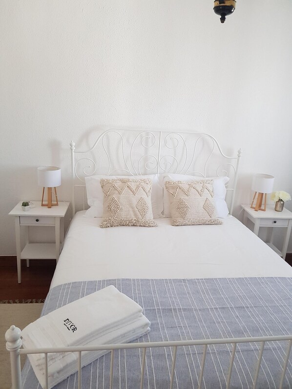 Un lit bien fait, avec une tête de lit en métal blanche, deux coussins décoratifs, une couverture à rayures et une serviette pliée, le tout posé sur un cadre de lit en métal.