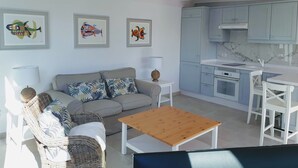 Een moderne woonkamer met een bank, salontafel en rieten stoelen. Grenzend hieraan bevindt zich een keuken met lichtblauwe kastjes en een wit aanrecht.