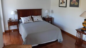 Een keurig opgemaakt slaapkamer met een bed, twee nachtkastjes met lampen, een klein tafeltje met een lamp, en twee ingelijste kunstwerken aan de muur.