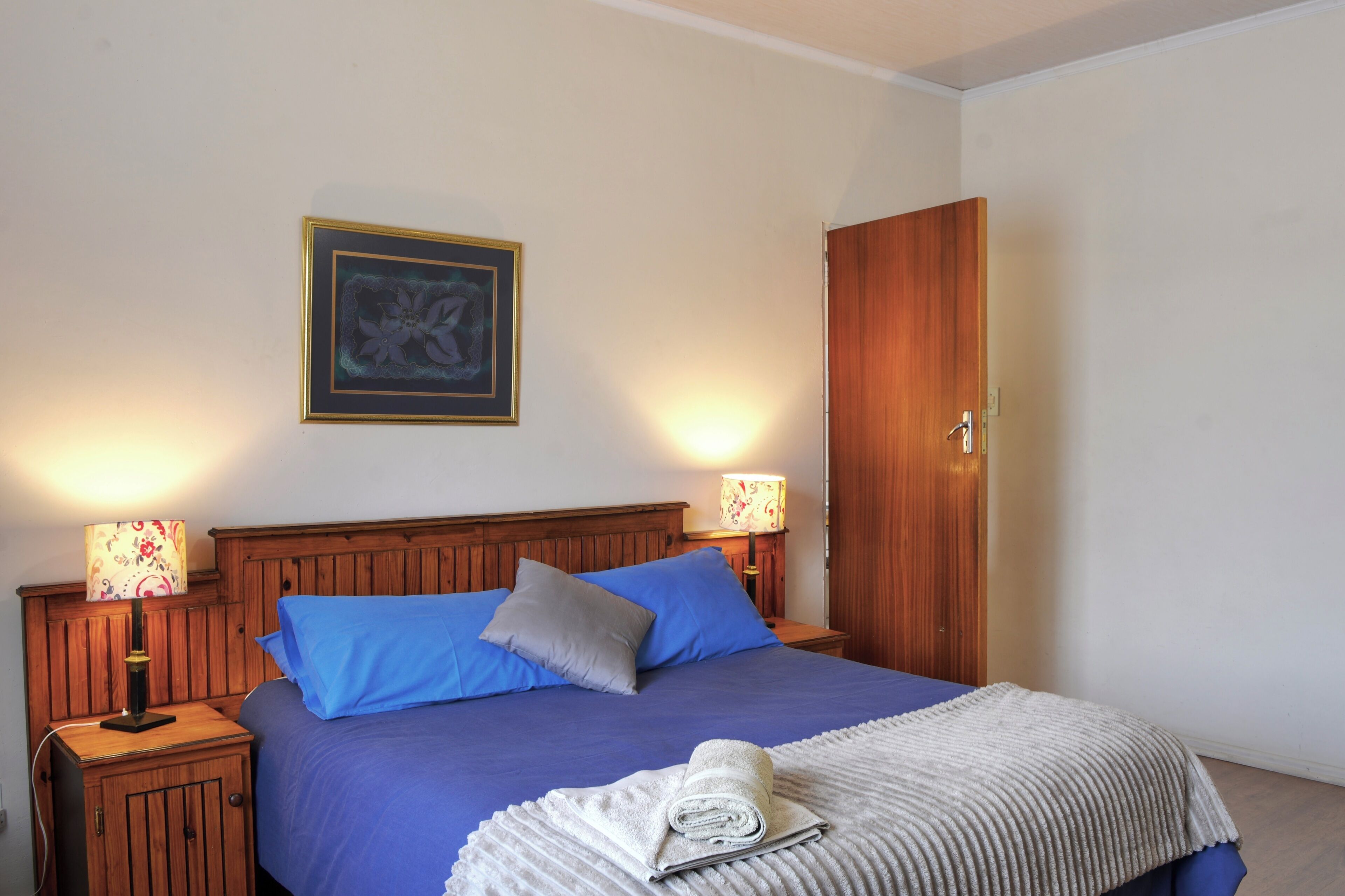 Cama bem arrumada com almofadas azuis e cinzas, cabeceira de madeira e uma obra de arte emoldurada acima.