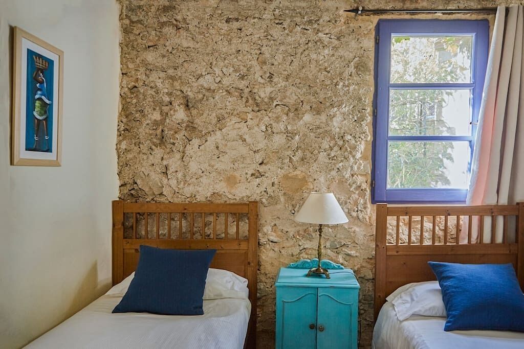 Ein Zimmer mit zwei hölzernen Betten, einer Steinwand, einem Fenster mit blauen Rahmen und einem kleinen, blaugrünen Schrank mit Lampe.