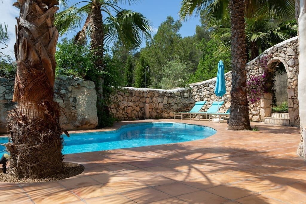 A swimming pool with stone walls and palm trees, surrounded by lounge chairs and a stone archway.