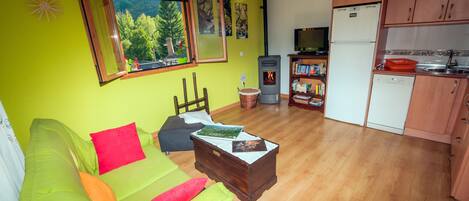 Un espacio habitable compacto con un sofá verde, una mesa de centro de madera, una zona de cocina con armarios de madera, un refrigerador y una ventana con vistas a árboles.