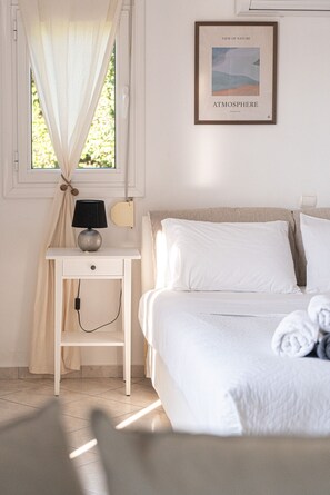 A bedroom with a bed, a bedside table with a lamp, a framed picture on the wall, and a window with curtains. - Charmful villa, stunning views,pool, hot tub & BBQ (Roussospiti)