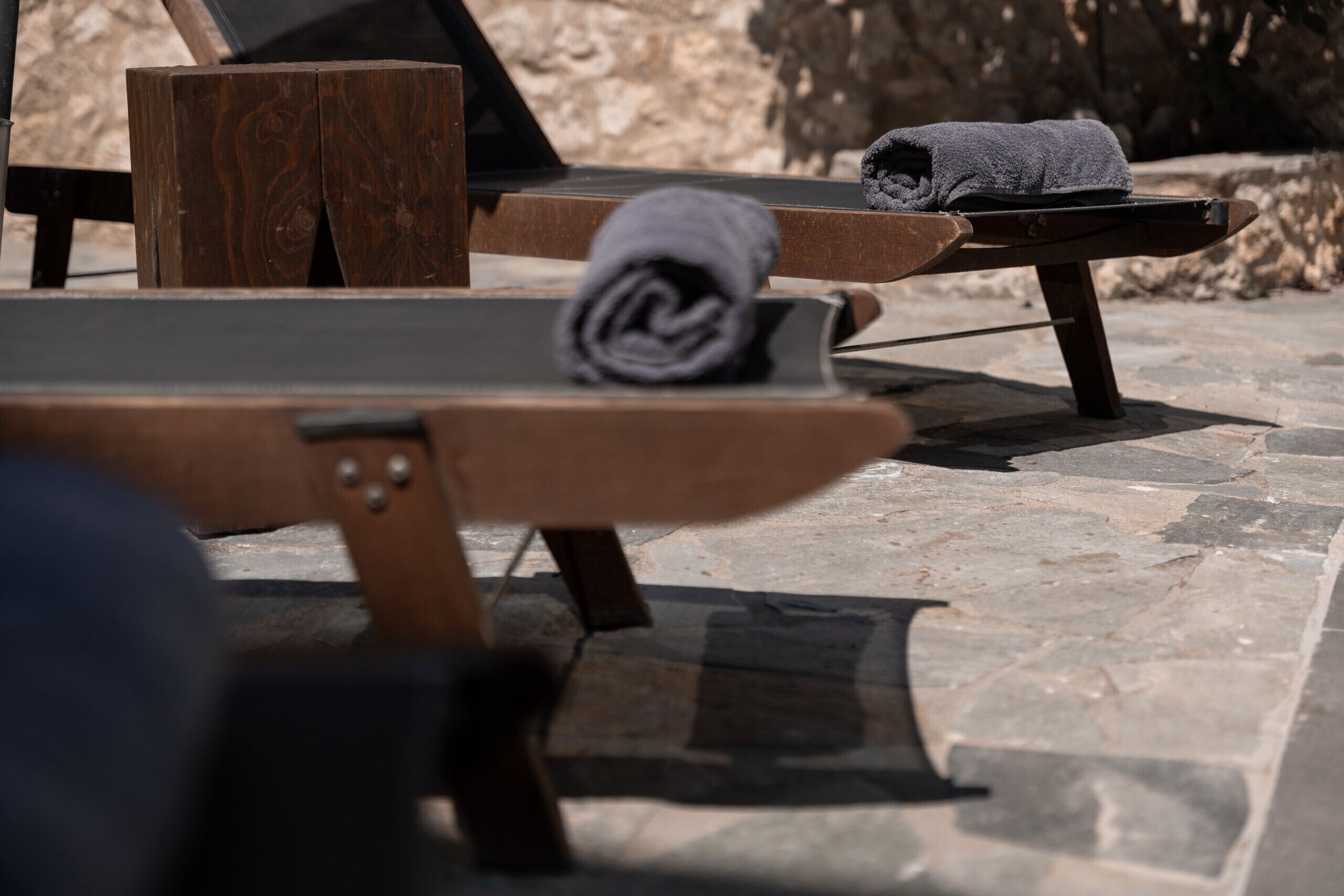 A stone-tiled outdoor seating area with wooden lounge chairs and a rolled-up towel on one of the chairs.