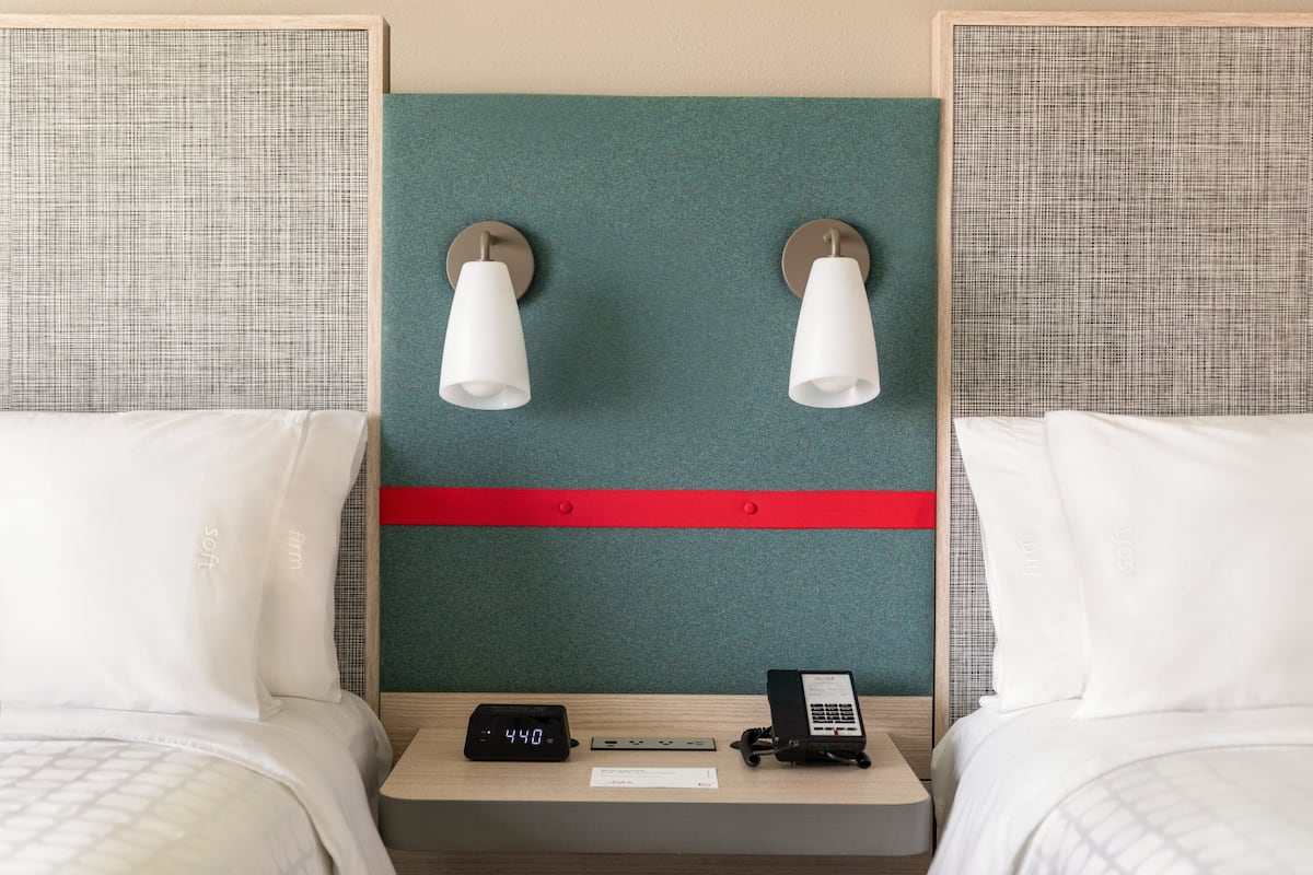 Ein Hotelzimmer mit zwei Betten, einem Nachttisch mit digitaler Uhr, einem Telefon und Wandleuchten.