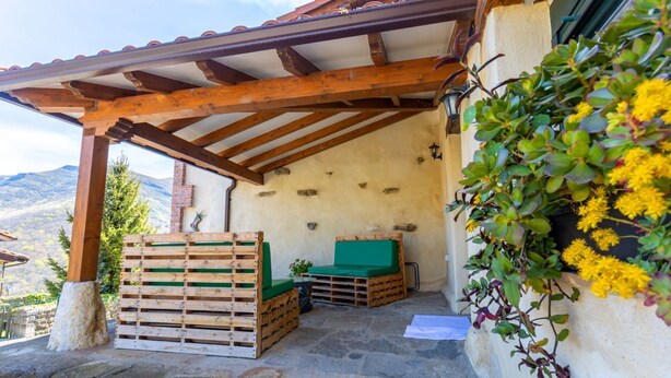 Un espace extérieur abrité, doté de bancs en bois et d’un coussin vert, d’un mur en pierre et de fleurs jaunes.