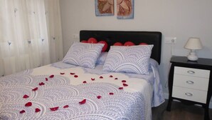 Un lit bien fait, avec une literie à motifs et des oreillers rouges, une table de chevet avec une lampe, et un mur orné de quatre œuvres d’art encadrées.