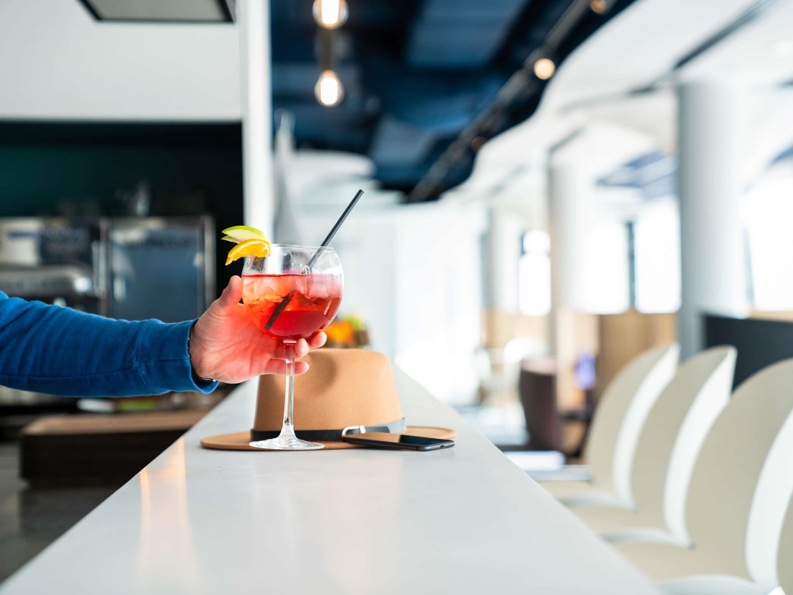 A hand holding a cocktail with a straw and garnish, placed on a table in a modern interior with a bar area in the background.