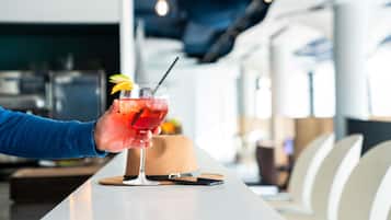 Una mano sosteniendo un cóctel con pajita y decoración, sobre una mesa en un interior moderno con una zona de bar al fondo.